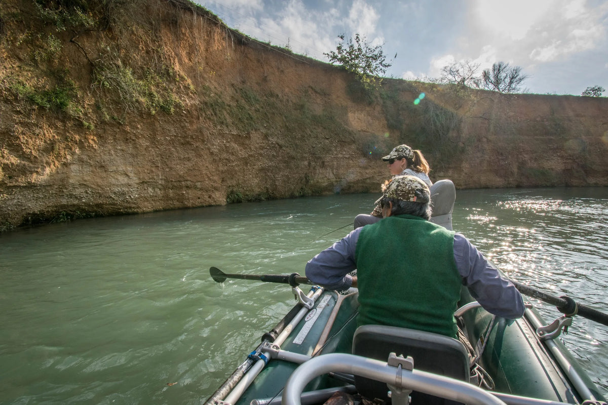 Marcus Rodriguez of Living Waters Fly Fishing rowing in a small inflatable boat on calm greenish water, with a steep sandy cliff wall rising on both sides under a partly cloudy sky.