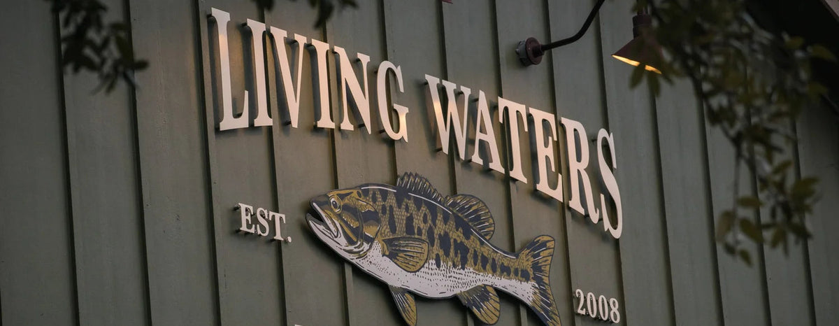 A wooden sign on a green siding wall reads “LIVING WATERS” and “FLY FISHING,” with a large Guadalupe Bass illustration in the center and “EST. 2008” beside it.